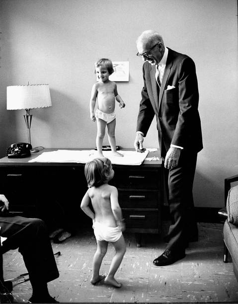 Pediatrician Dr. Benjamin Spock looking amused by two young patients during examination, c.1964