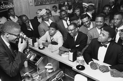 photo of then-Cassius Clay at lunch counter surrounded by fans with Malcolm X taking a picture in Miami, 1964
